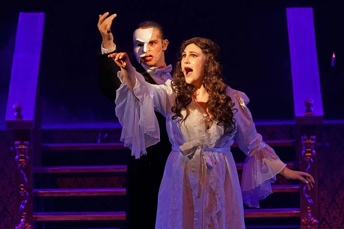 Two actors in a dramatic stage scene, one wears a half mask, both appear to be singing passionately.