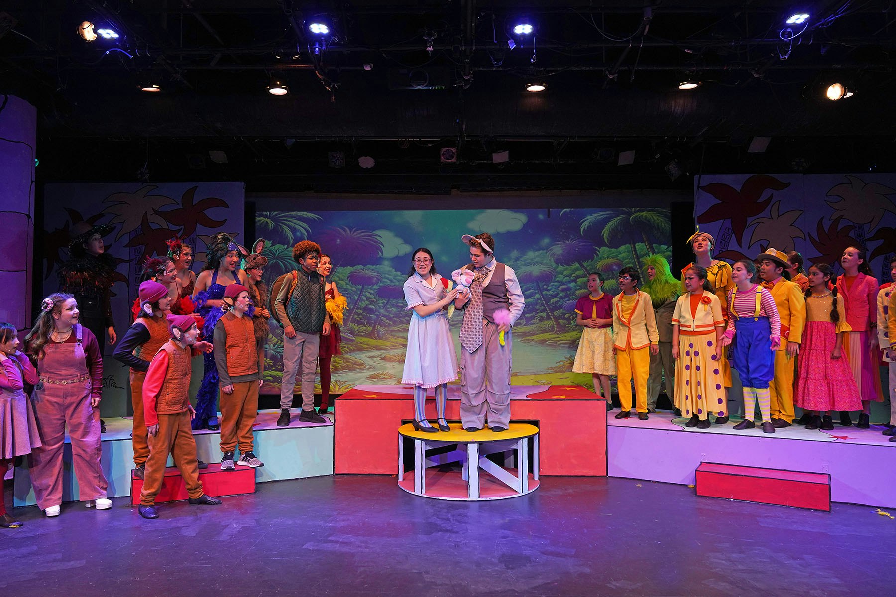 A lively stage performance featuring colorful costumes, with actors interacting and a whimsical backdrop.