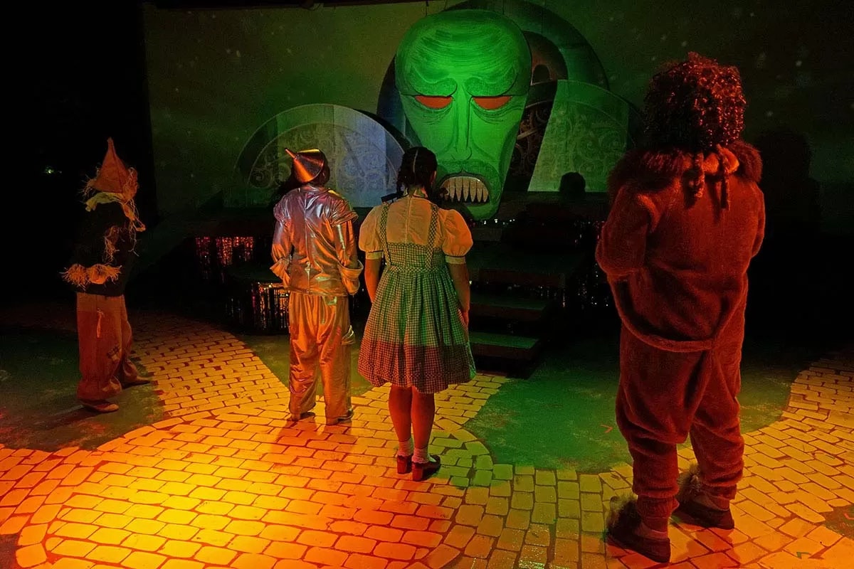 Four characters in costume face a large green face projection in a dark theater setting.