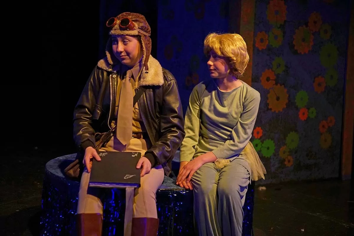 Two people in costume sit on stage, one dressed as an aviator and the other in a green outfit, against a colorful floral backdrop.