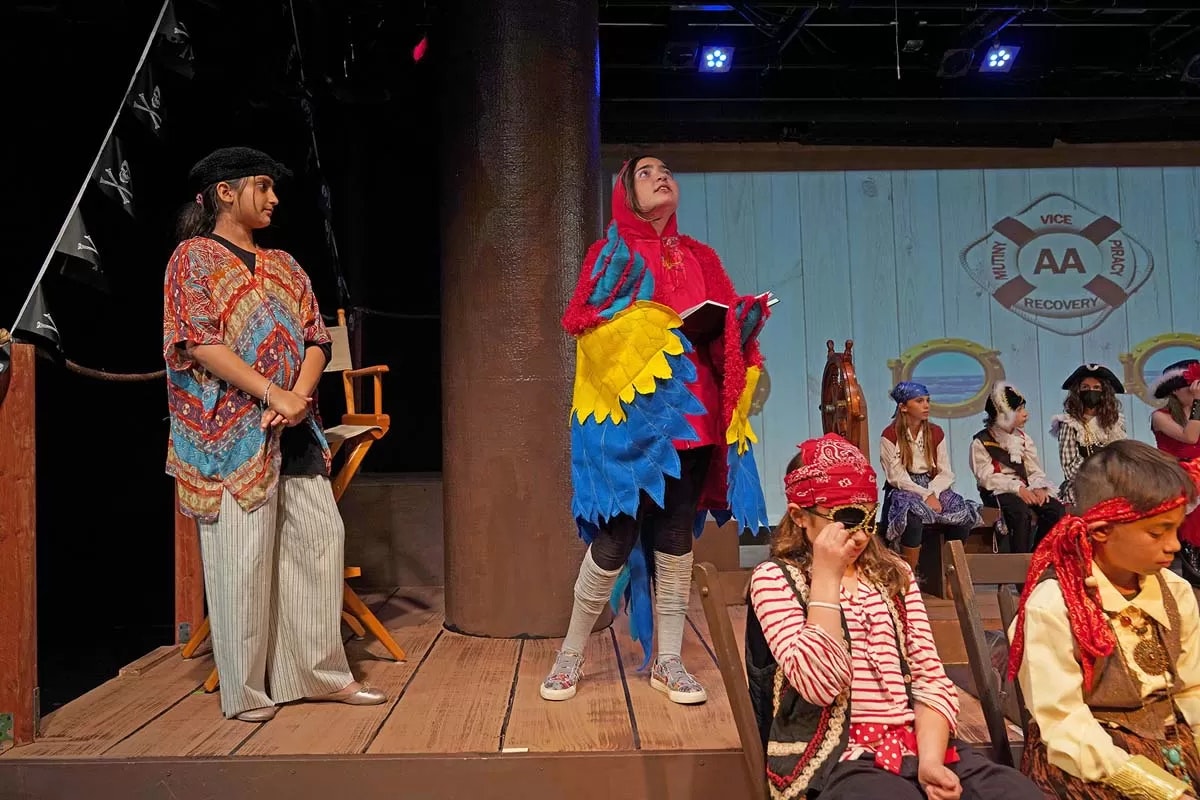 Kids in a play; one in a parrot costume, others dressed as pirates on stage.