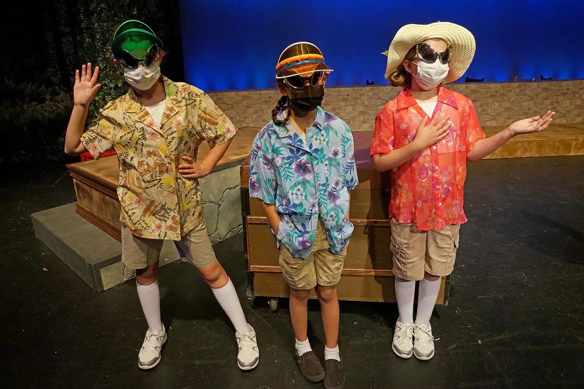 Three people in colorful shirts, shorts, socks, and masks posing on stage with props.