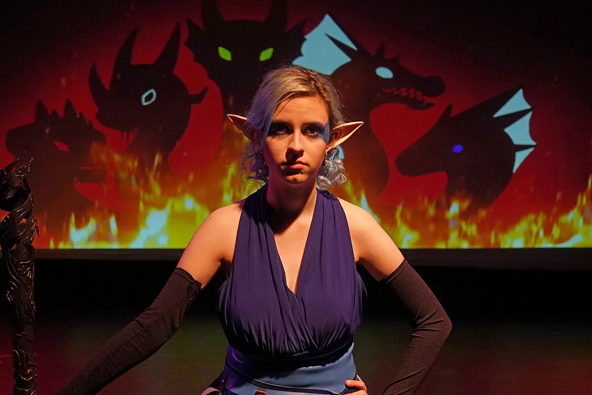 A person in fantasy costume with elf ears stands before a backdrop of fiery silhouetted creatures.