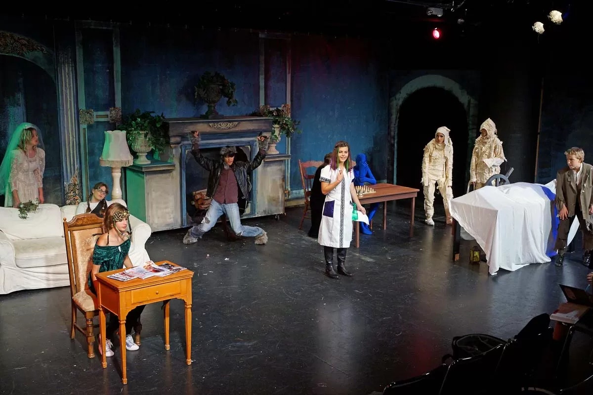A stage play with actors in various costumes, including a nurse, mummies, a person in blue, and a bride, set in a room with a sofa and table.