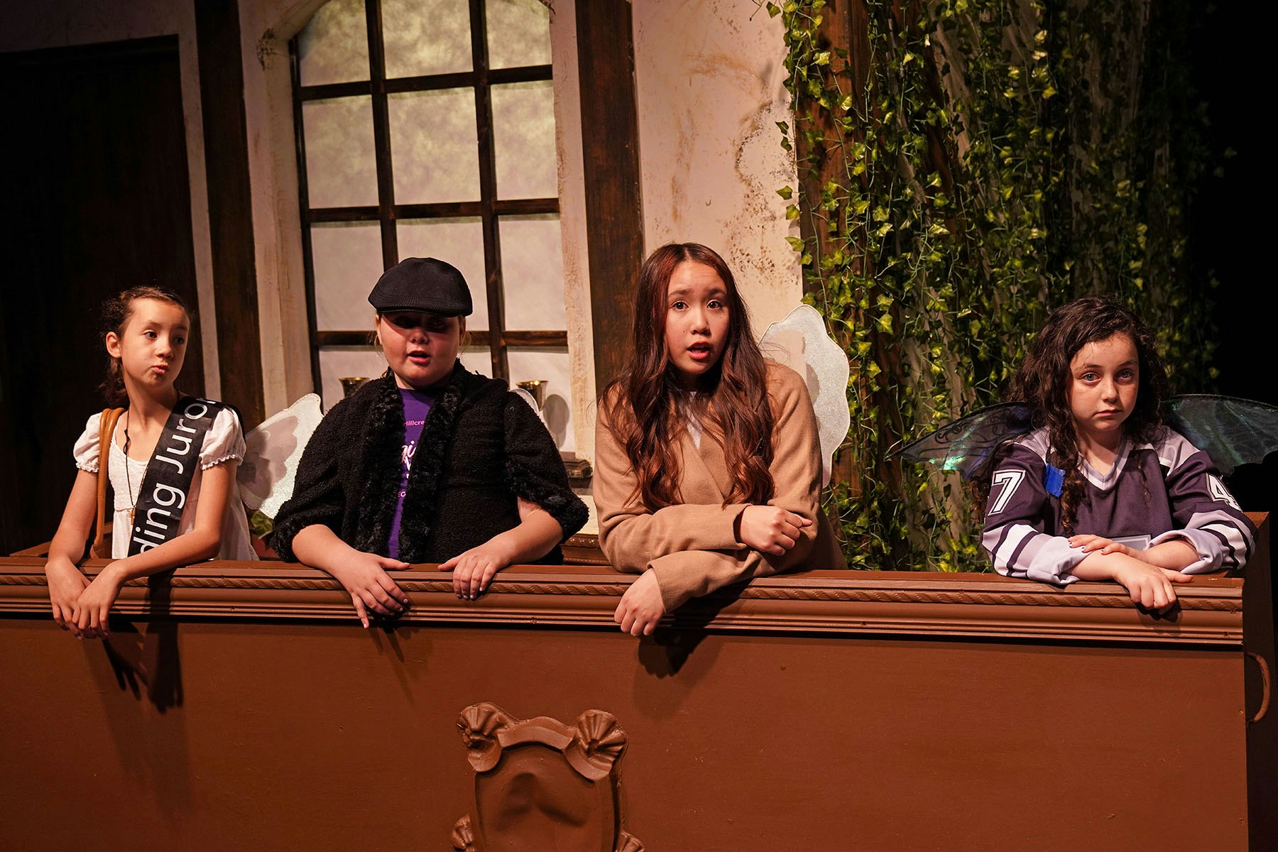 Four people in costume stand behind a wooden box, with a window and ivy backdrop.