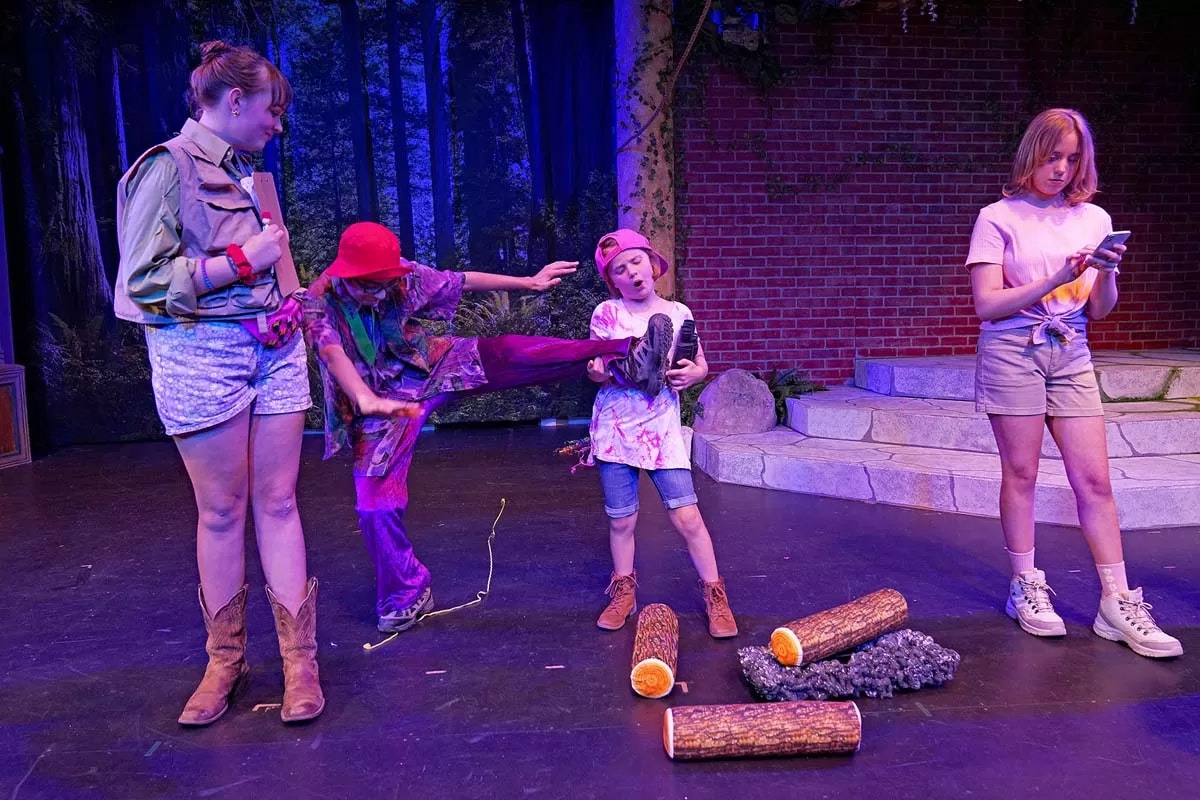 Four people on stage, one kicking another, surrounded by faux logs, against a forest and brick backdrop.