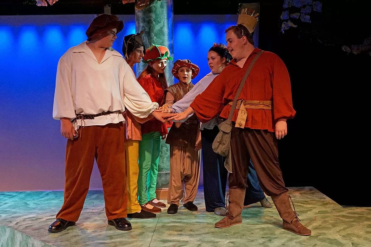 A group of people in colorful costumes stand on a stage with hands together in the center.