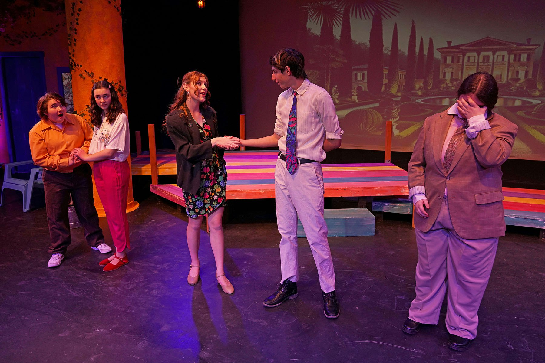 Five people on stage in colorful attire, performing a play with a painted set and backdrop.