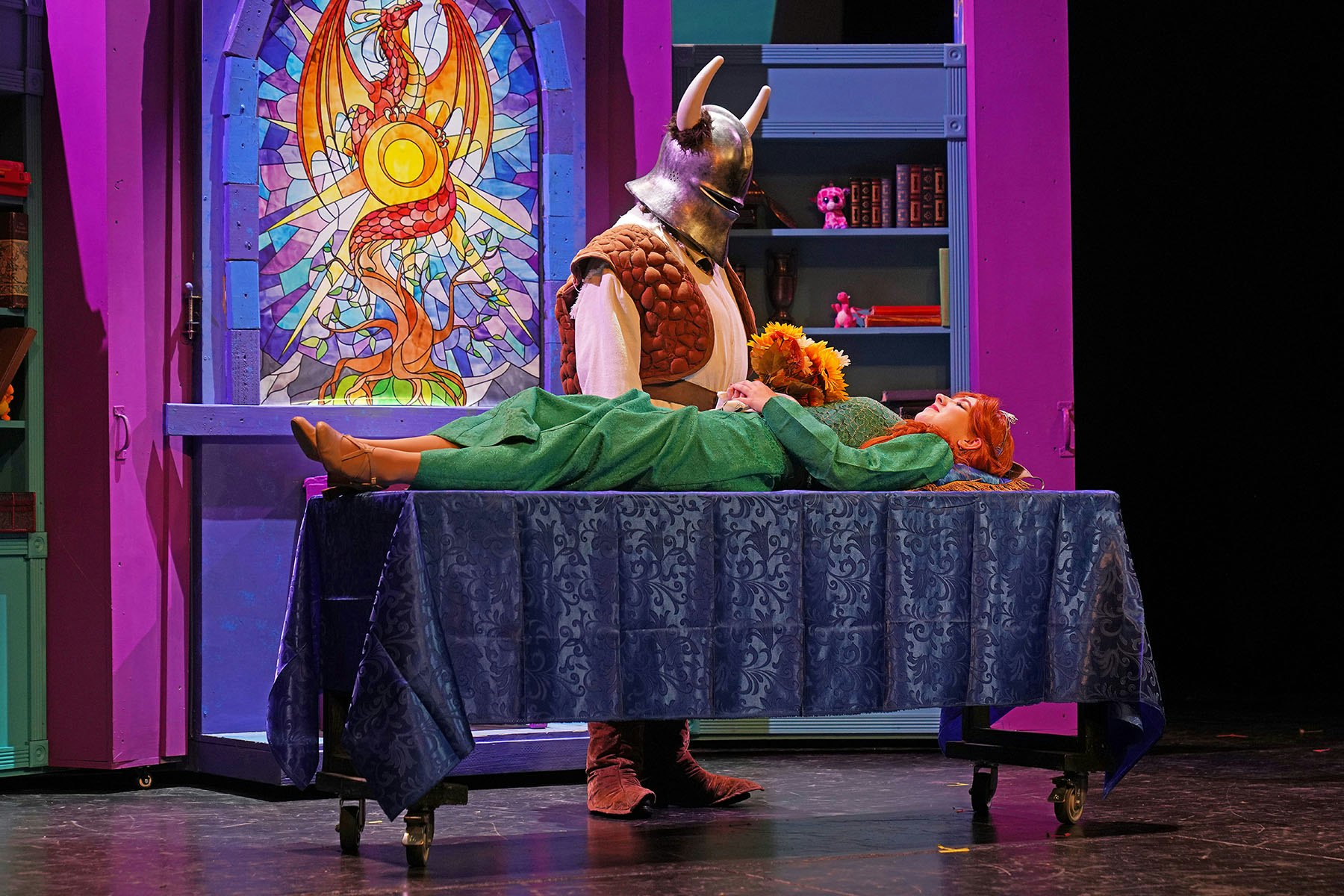 A person in armor stands by a woman in green on a table, with a stained glass dragon window backdrop.
