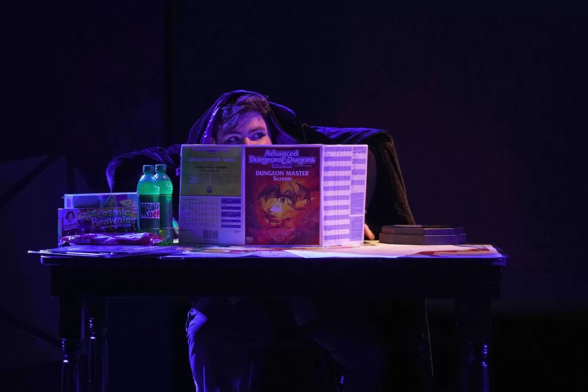 A person in a hood at a table with a Dungeons & Dragons screen, snacks, and soda.