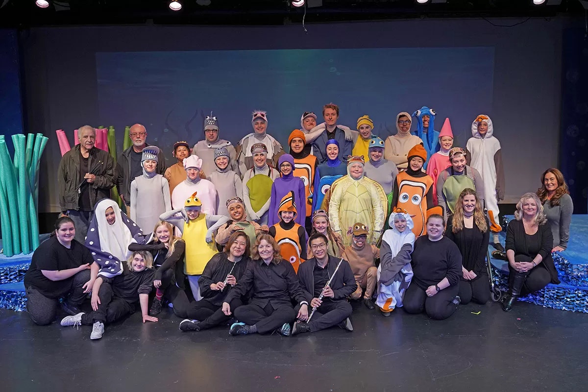A group in ocean-themed costumes poses on stage, with musicians in black seated in front.