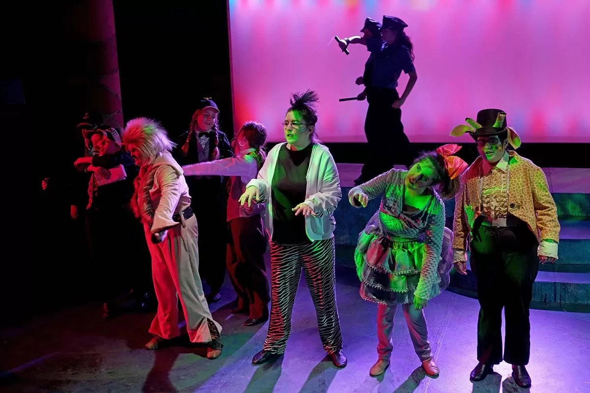 A group of performers in colorful, theatrical costumes standing on stage with vibrant lighting.