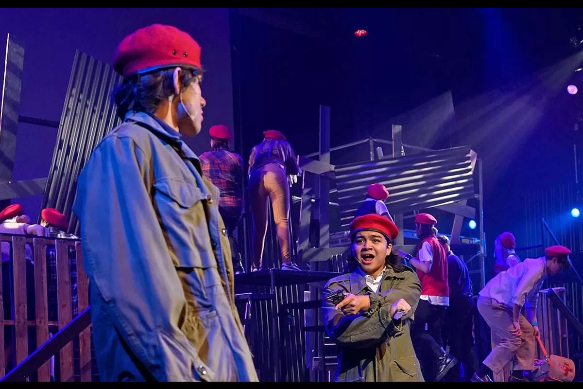 People on stage in red berets and costumes, performing a theatrical scene with dramatic lighting.