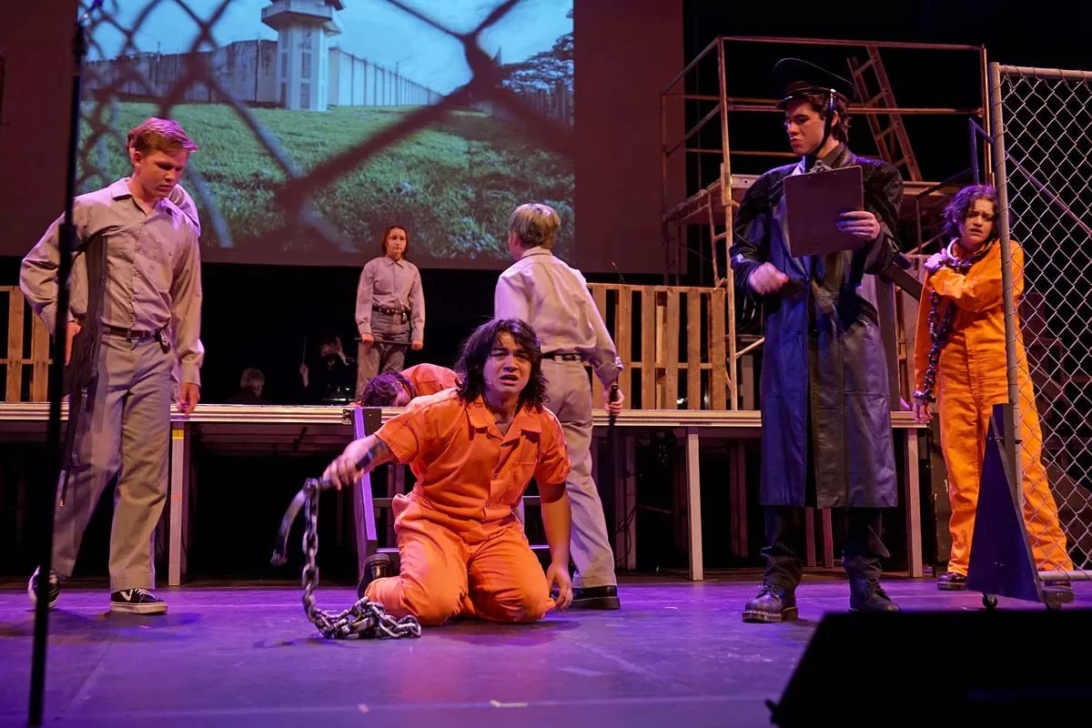 A theatrical scene with people in costumes, chains, and a projected prison background.
