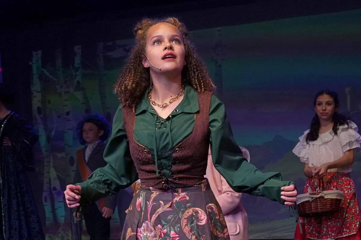 A theatrical performance with a young actor in a green shirt and floral skirt, looking upwards, two others in the background.