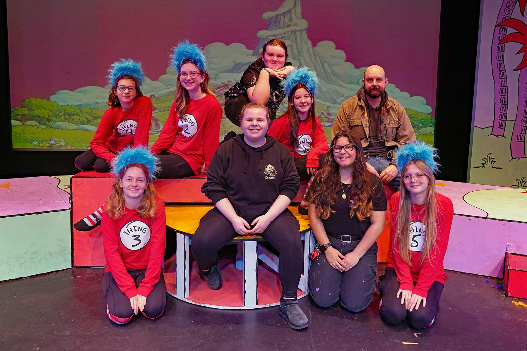 A group of performers dressed as "Things" from Dr. Seuss, posing on stage with colorful set pieces in the background.