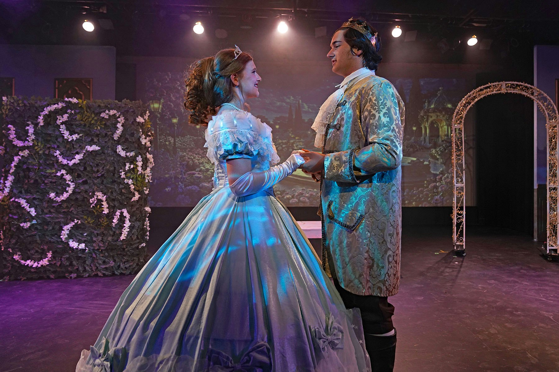 A couple in elegant costumes performs on stage, surrounded by decorative foliage and soft lighting, conveying a romantic atmosphere.