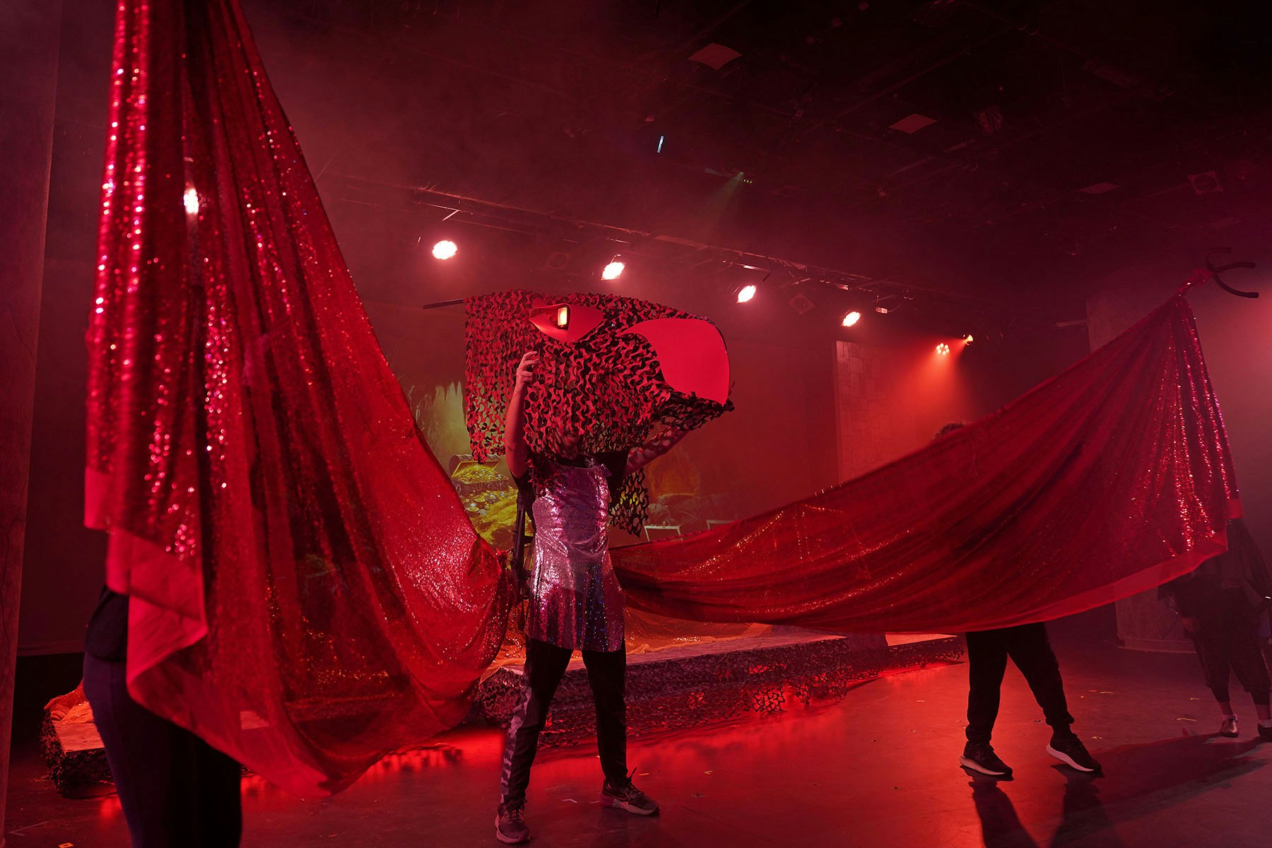 Performers holding red, shimmery fabrics on stage, with dramatic lighting and props in the background.