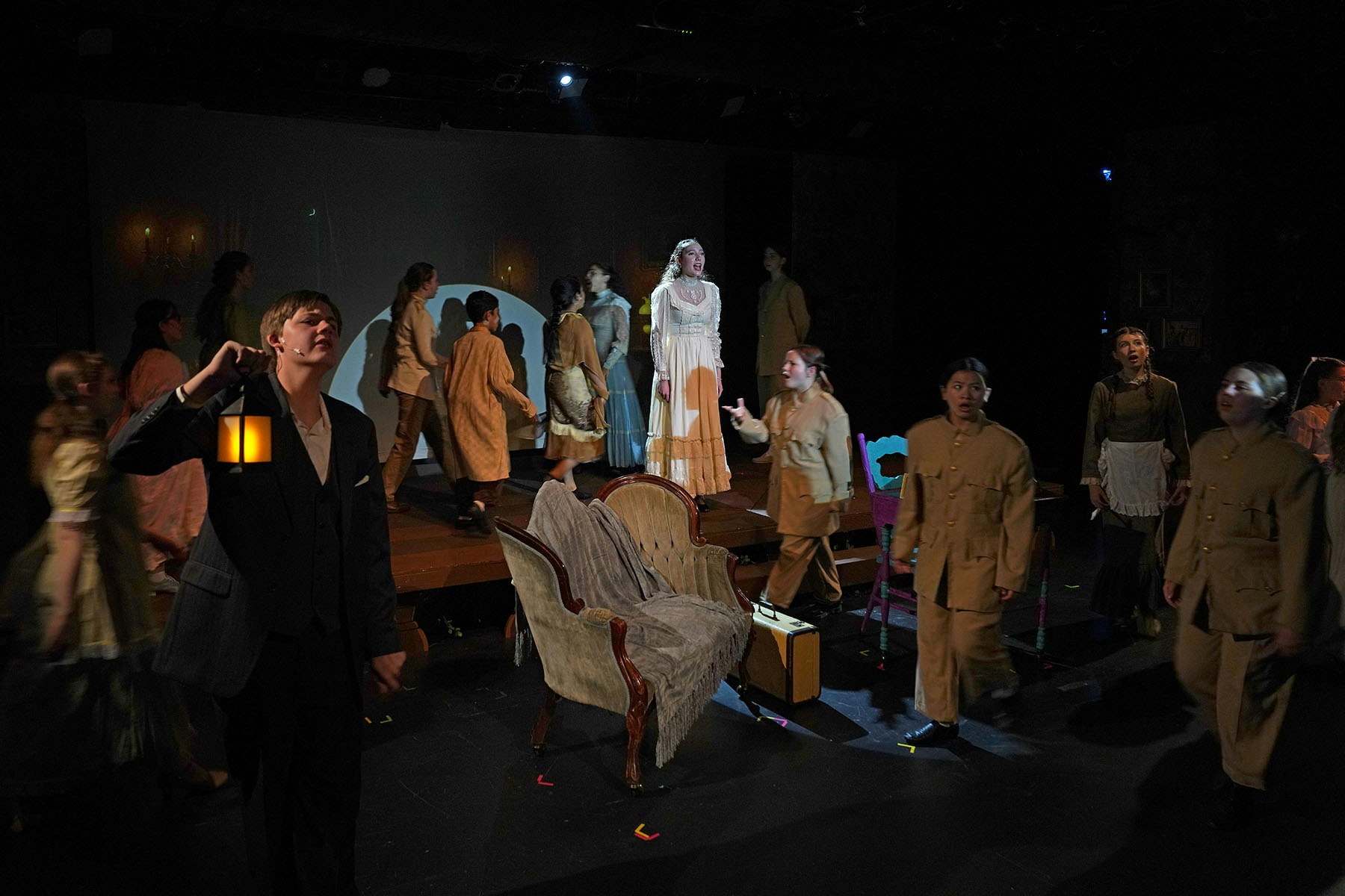 A theatrical performance with actors in period costumes on stage, including a person holding a lantern.