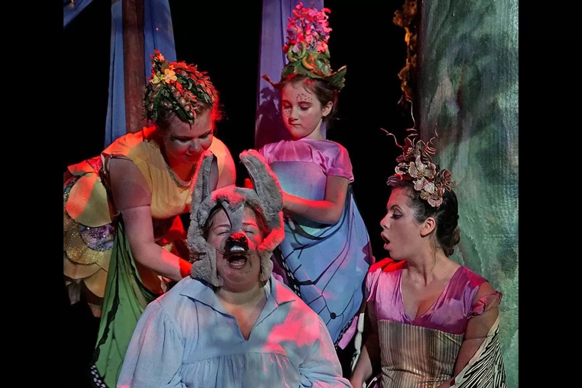 A theatrical scene with four performers, one wearing donkey ears, in colorful costumes and floral headpieces.