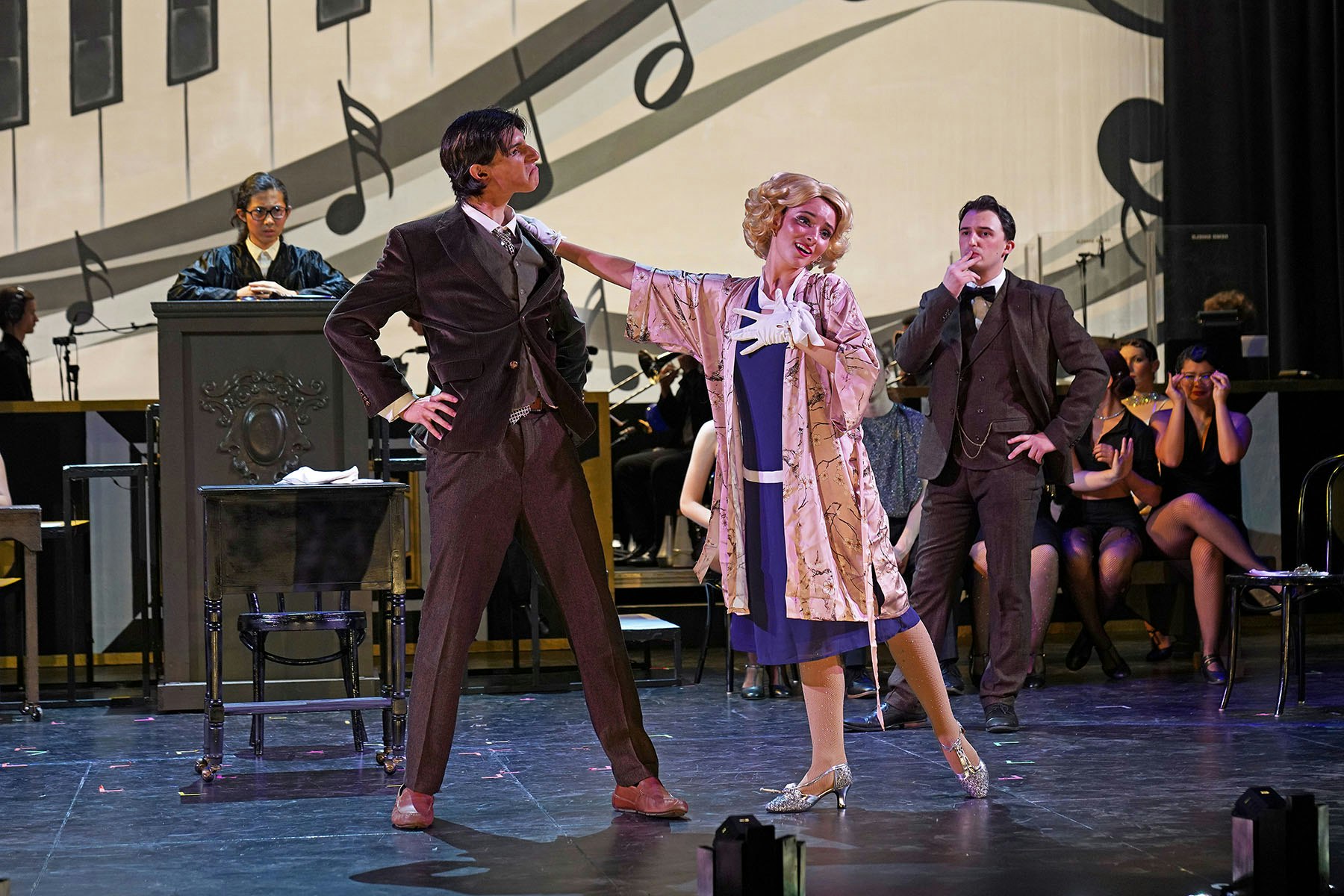 A theatrical performance featuring actors in vintage attire, showcasing dramatic poses on stage with a musical backdrop.