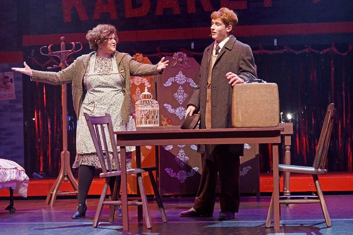Two actors on stage in a play; a woman gestures, a man holds a suitcase and hat, with a table and chairs in the scene.