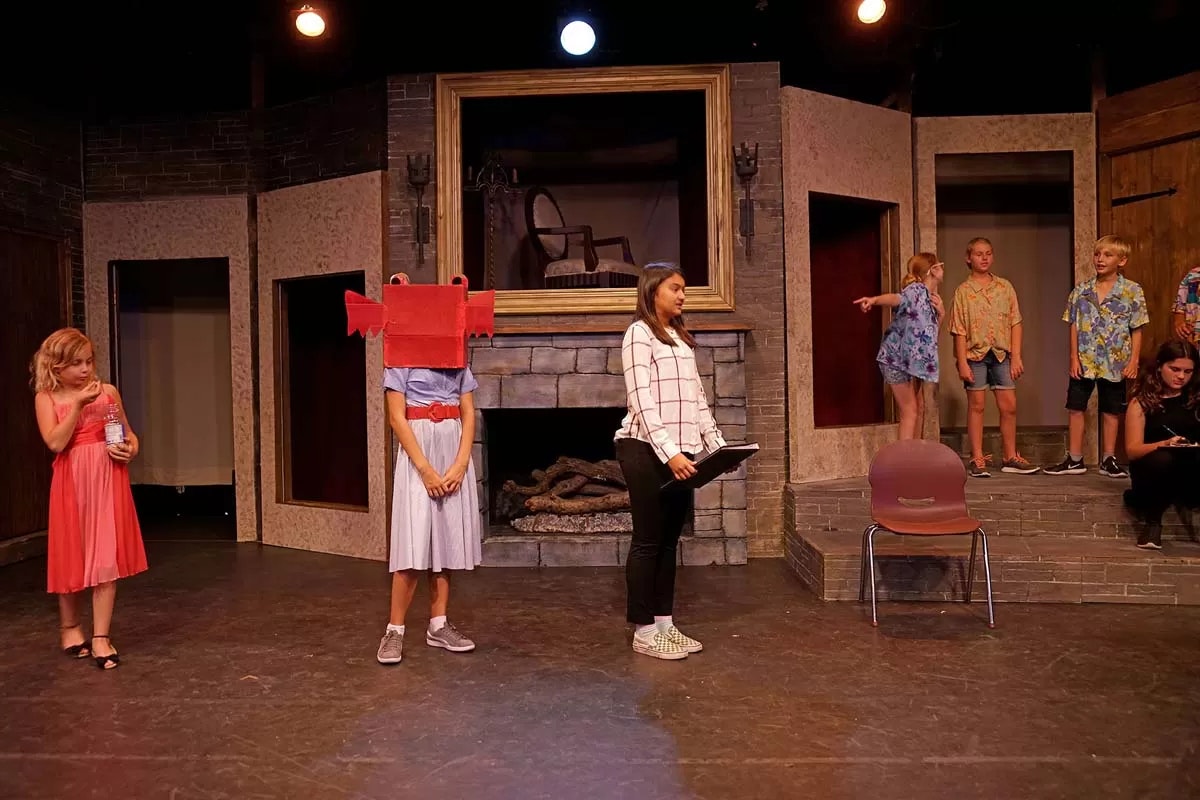 A stage play with children; one wears a red box, another drinks water. Clad in colorful outfits, they interact on a simple set.