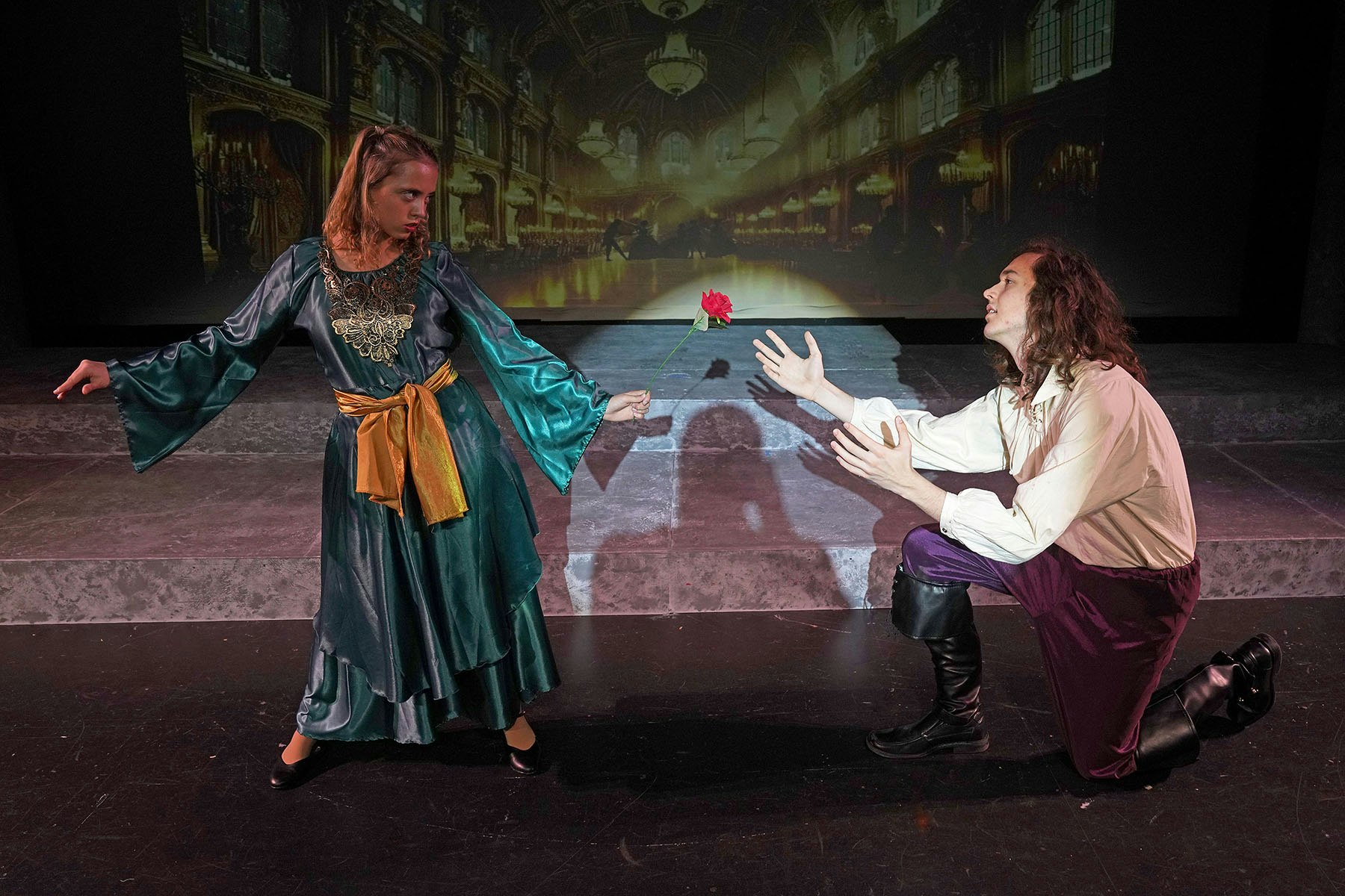 Two actors in a theatrical scene, one kneeling and reaching for a rose offered by the other, with a grand setting backdrop.