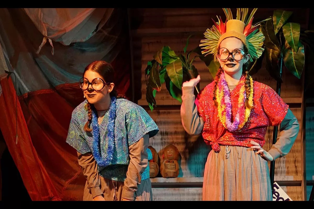 Two people in colorful outfits, big glasses, and face paint, with plants in the background, possibly performing on stage.
