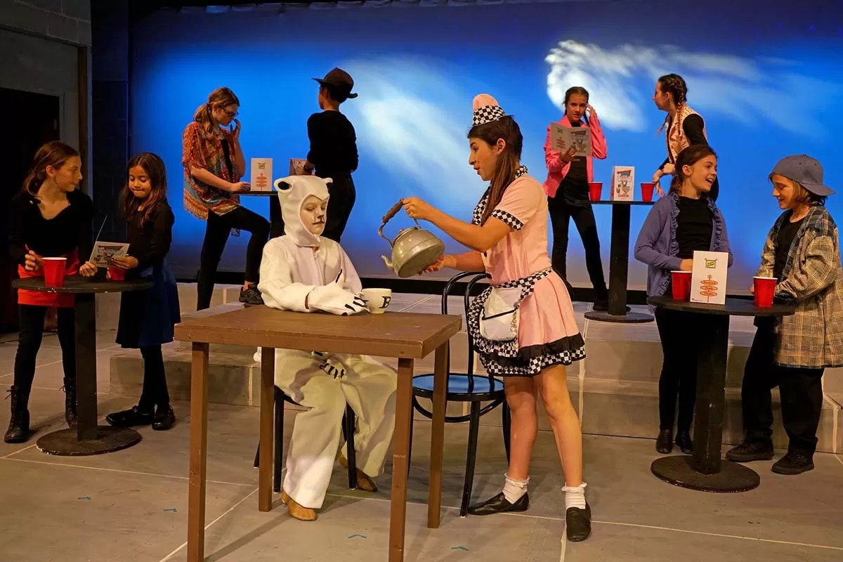 Children performing a play: one in a polar bear costume, others as diners and a waitress, with props like tables, menus, and a teapot.
