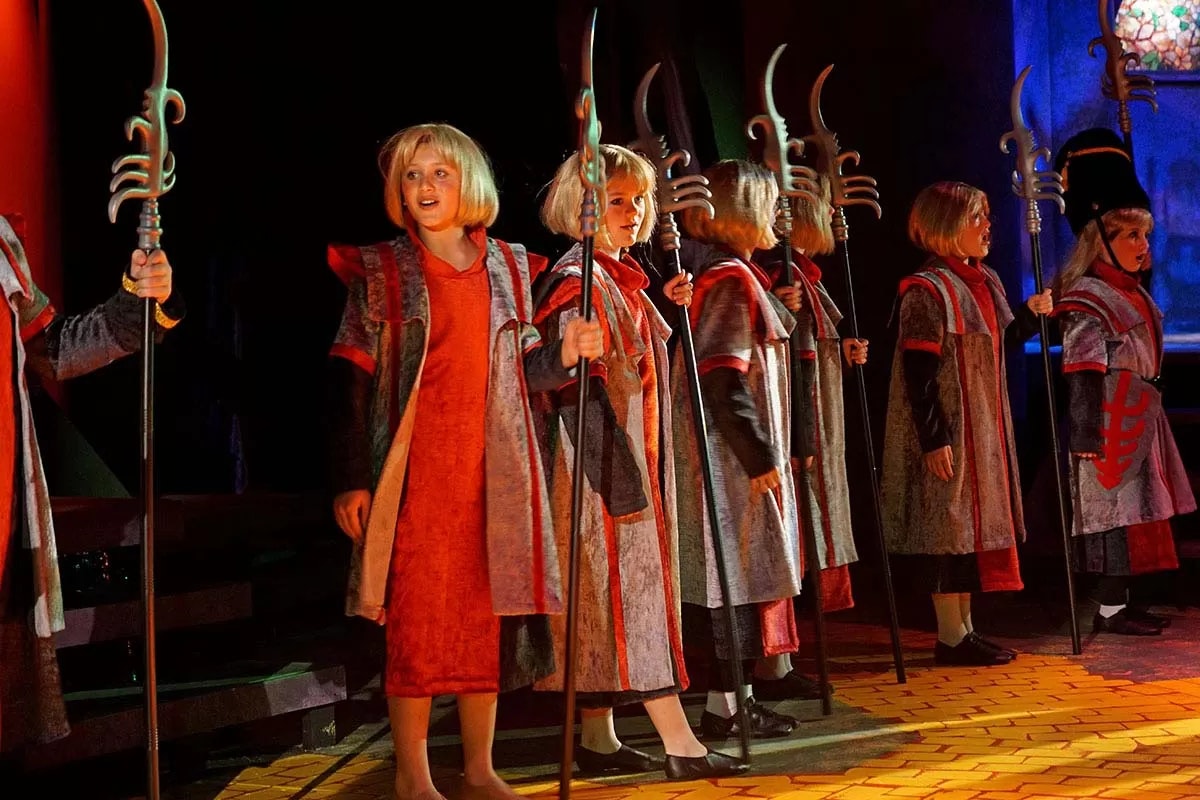 A group of people in costumes holding staffs, performing on stage with theatrical lighting.