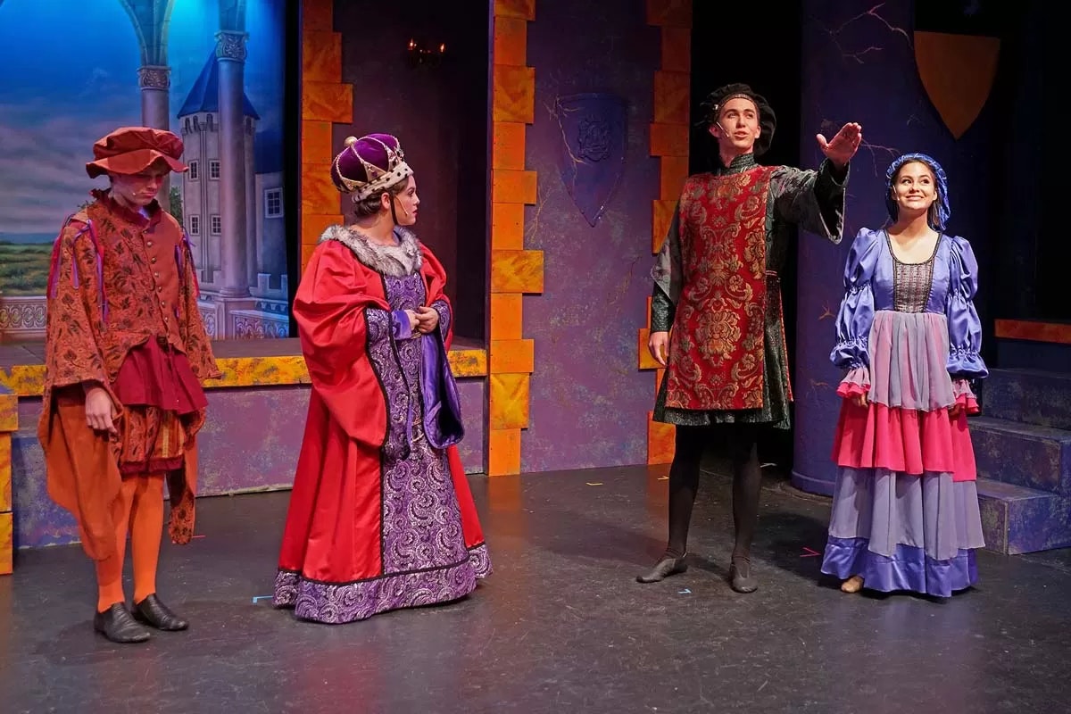 Four people in medieval costumes on stage with a castle backdrop.