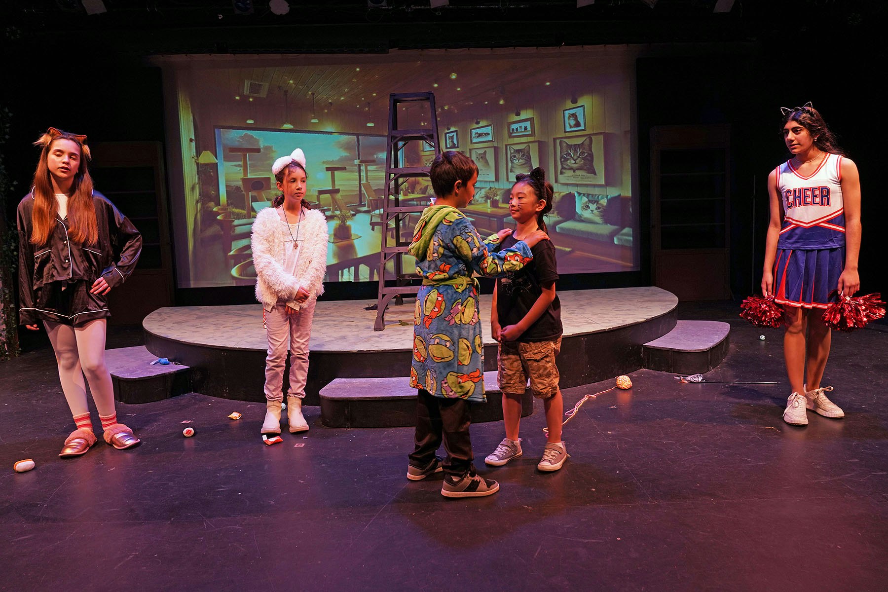 A group of kids in costumes are on a theater stage with a backdrop of a cozy room with cat images.