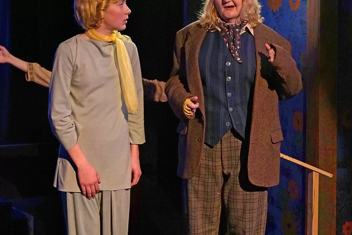 Two people in costumes on stage, one in a suit and tie, the other in a dress with a yellow scarf.