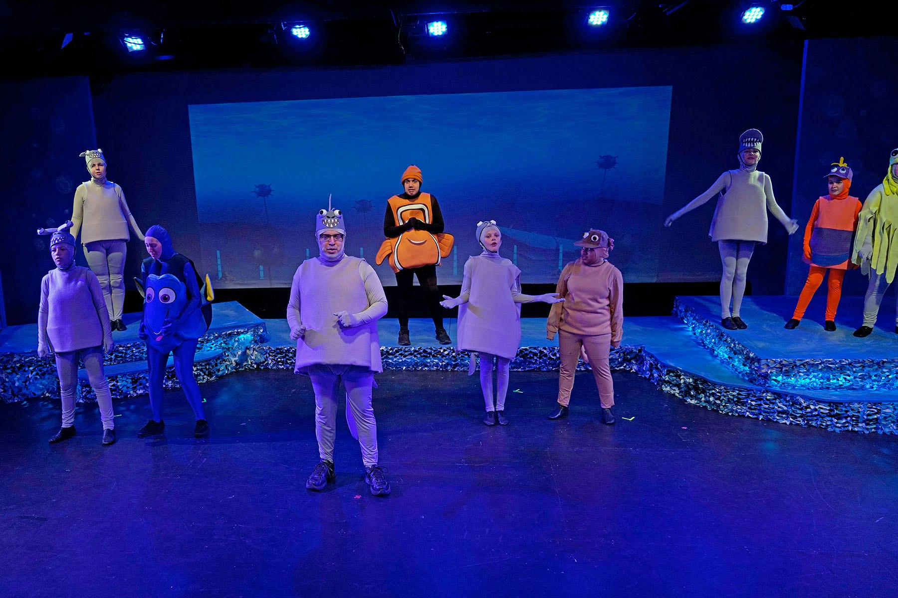 People in colorful fish costumes performing on a stage with blue lighting.