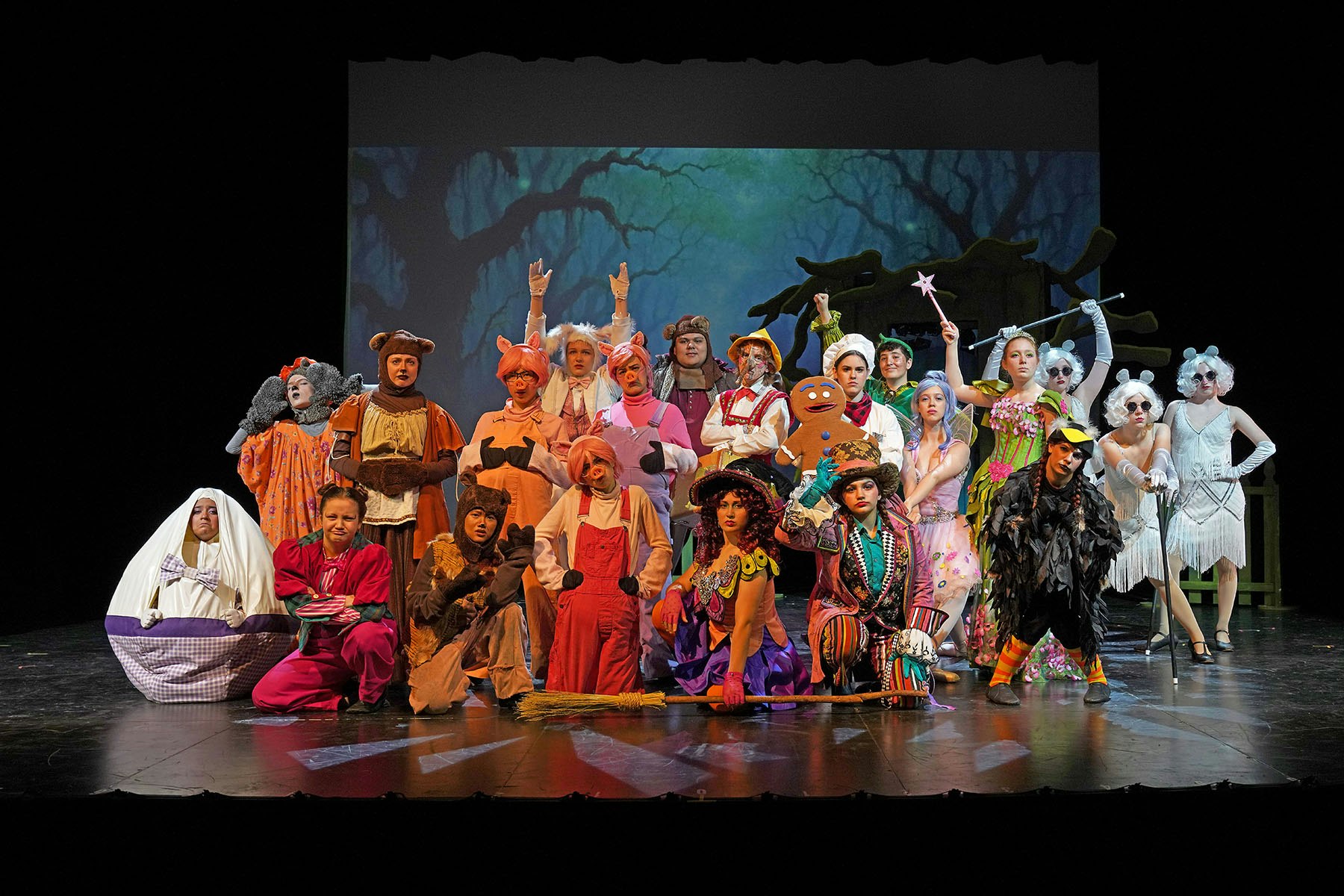 A group of people in colorful costumes pose on a stage with a forest backdrop.
