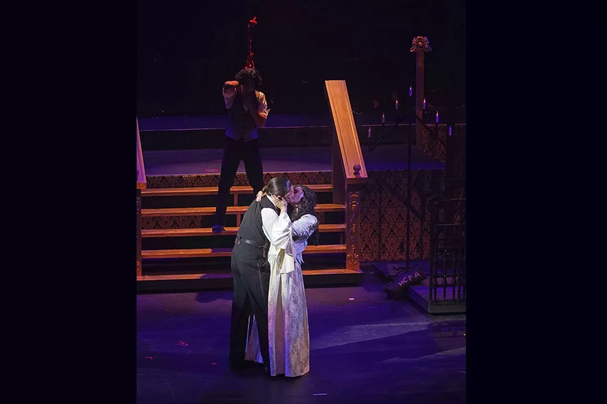 A theatrical scene with two people kissing on stage, another in the background covering his eyes.