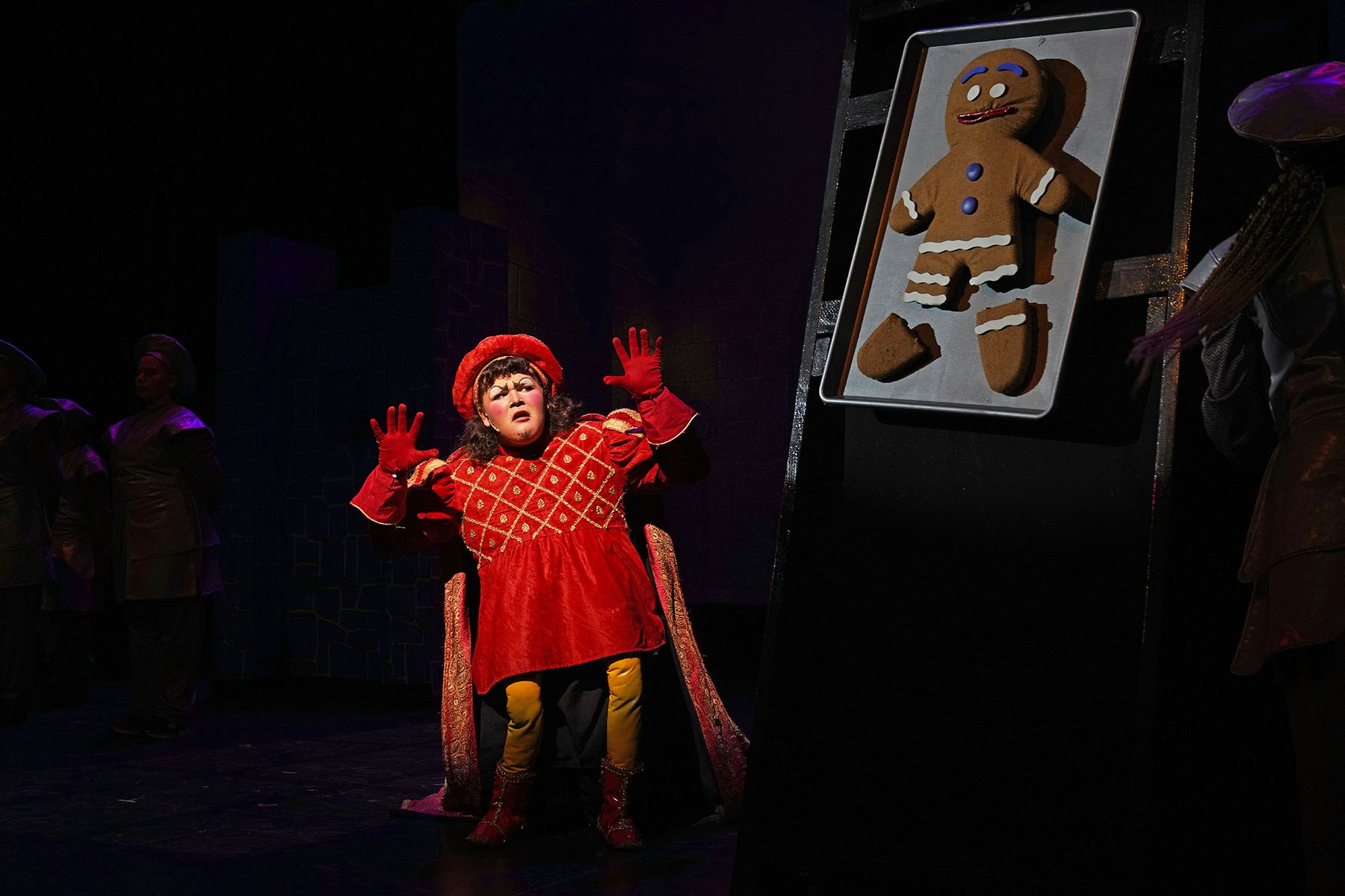 Person in red costume, surprised, with a large gingerbread figure on the right, on stage.