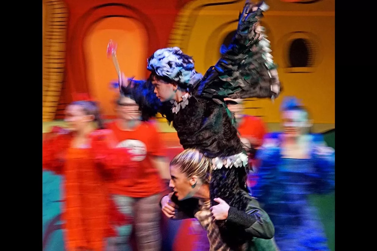Colorful stage performance with a person dressed as a bird on another's shoulders, surrounded by blurred motion of other performers.