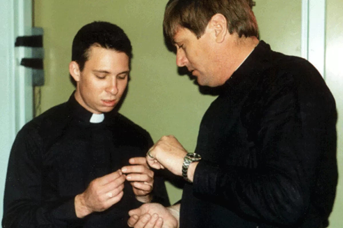 Two people in black clothing, one with a clerical collar, examining a small object in their hands.