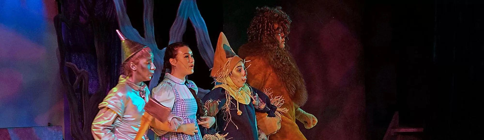 Four people in colorful costumes on stage, resembling characters from "The Wizard of Oz."