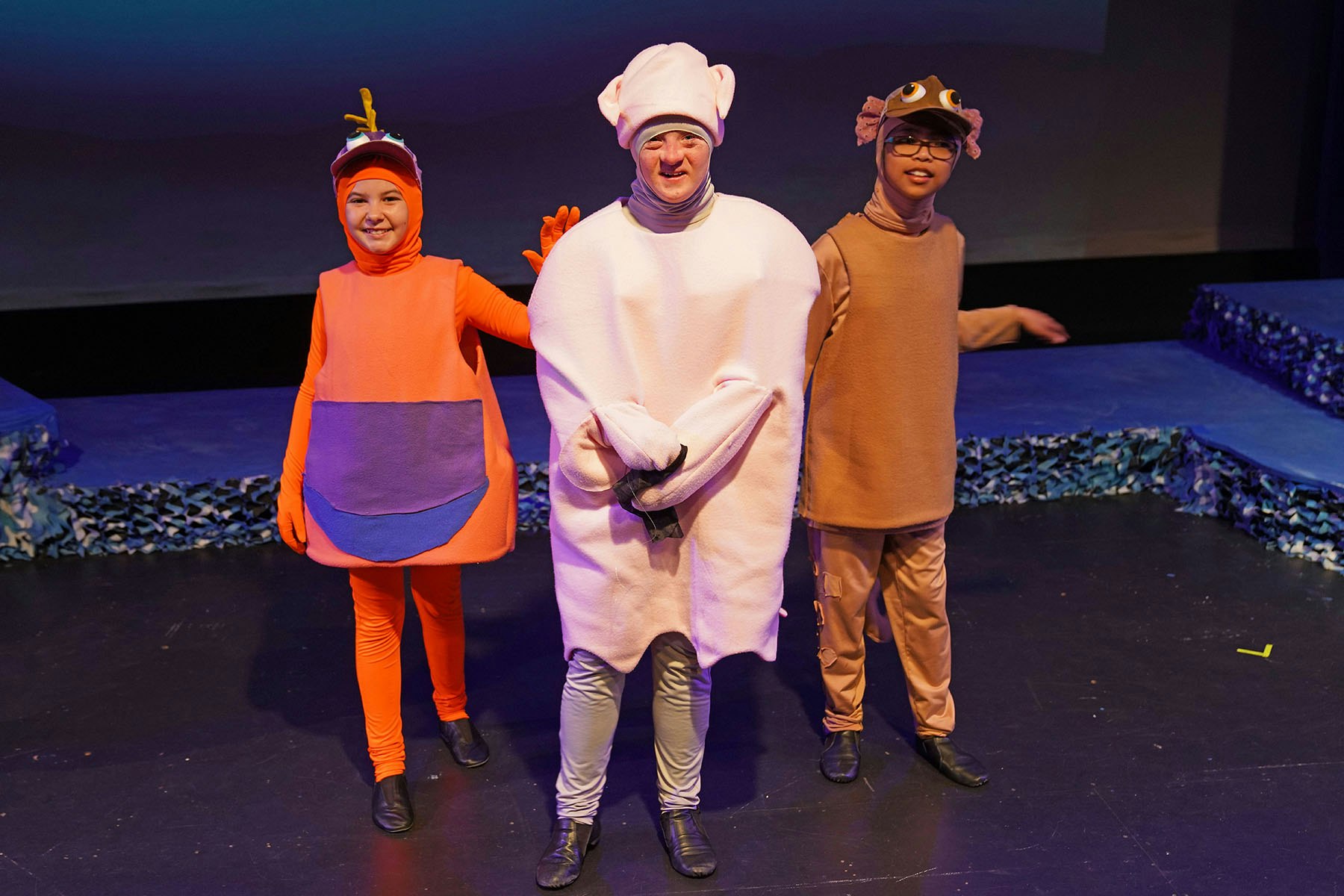 Three people in colorful, whimsical costumes on a stage, possibly for a performance or play.