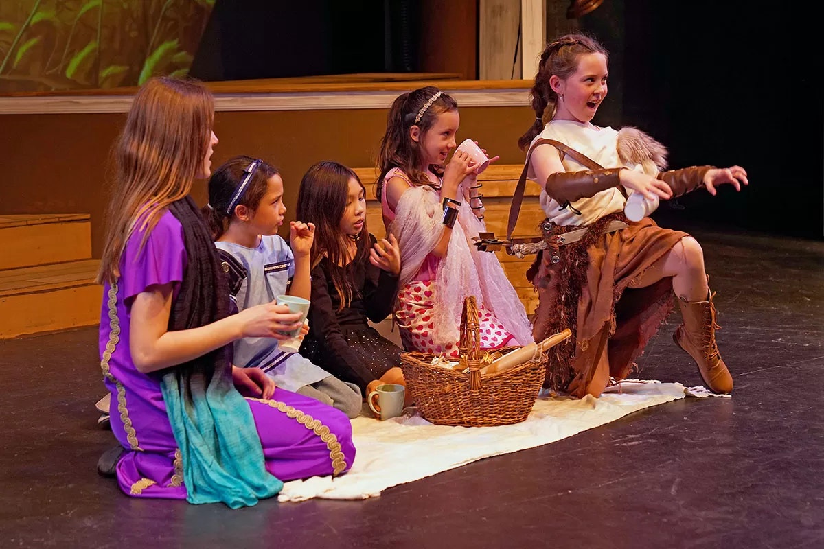 Five children in costumes doing a play, sitting on stage with drinks and a basket.