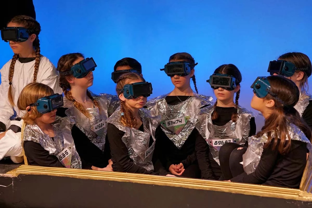 People wearing futuristic goggles and silver outfits are seated together, likely in a performance or sci-fi setup.