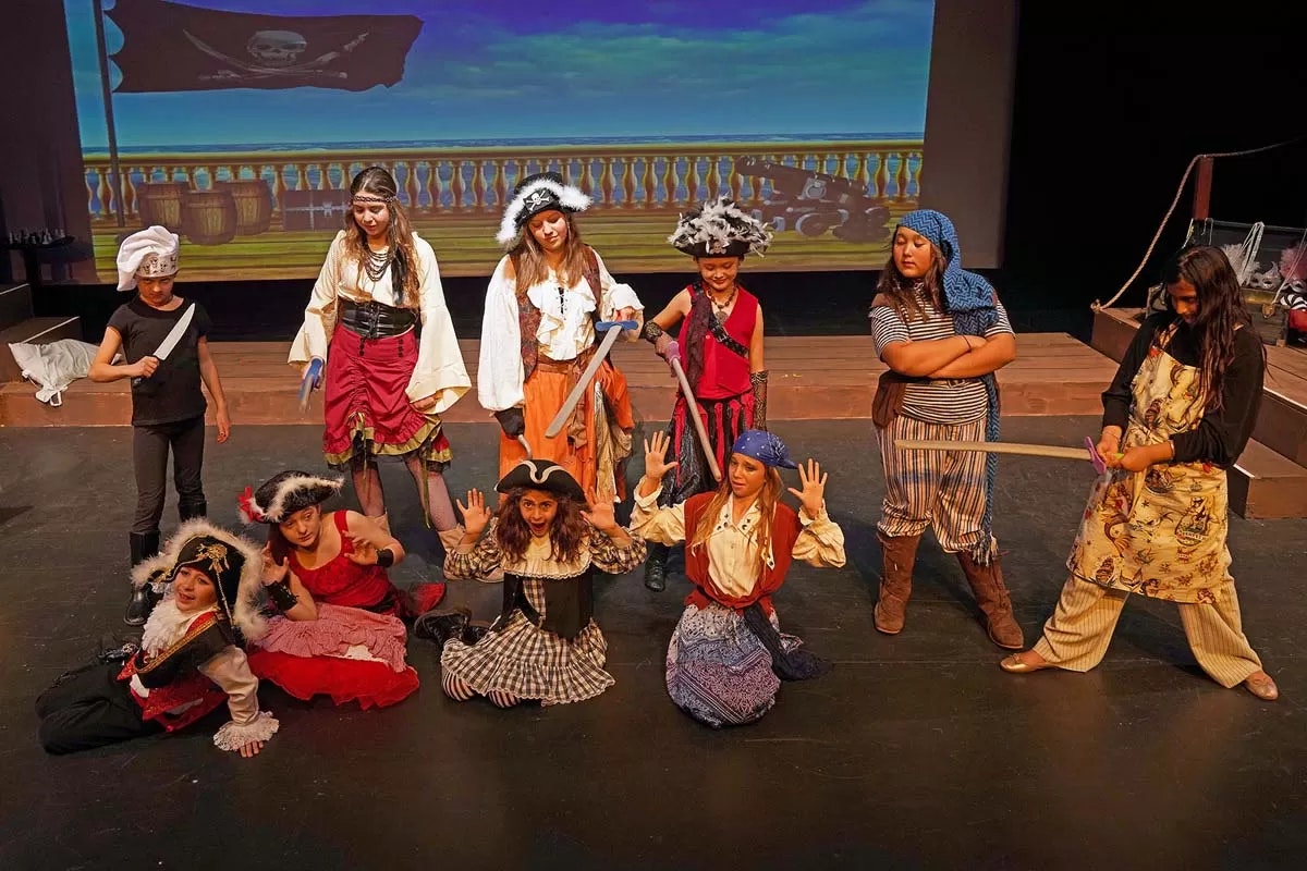 A group of kids dressed as pirates on stage, acting out a scene with props and a pirate ship backdrop.