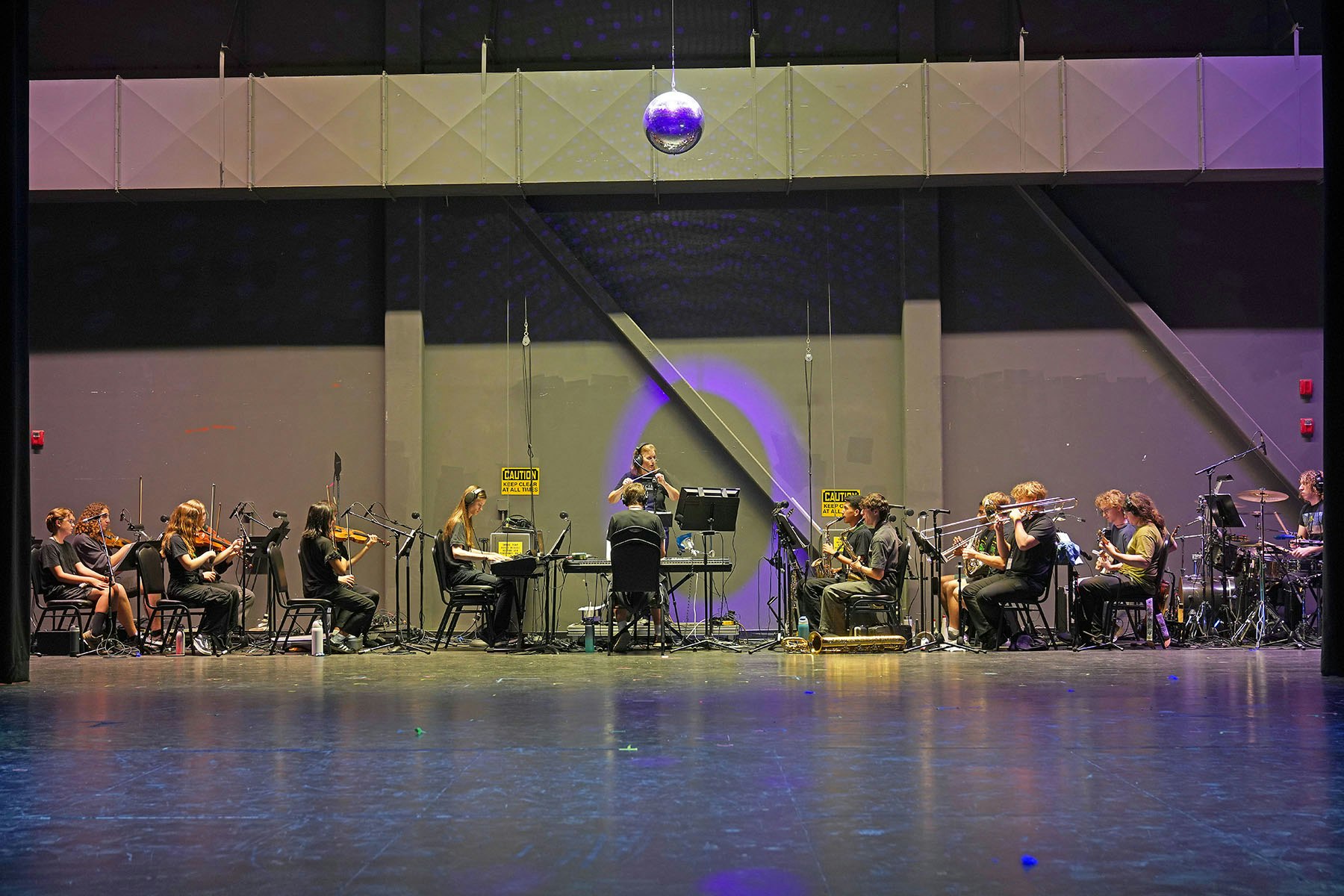 Orchestra rehearsing on stage under a disco ball with musicians playing instruments and a conductor in the center.