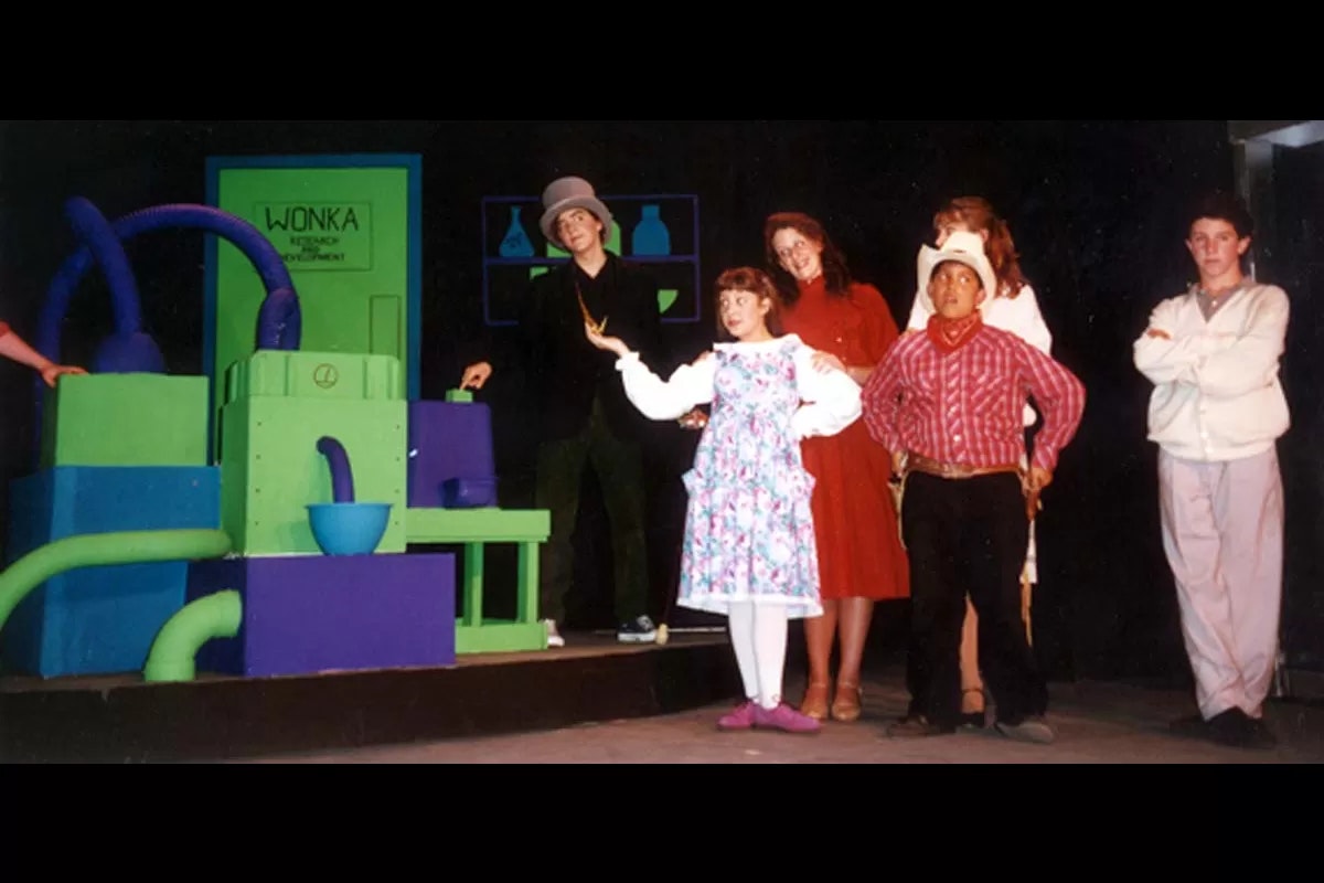 A group of young actors in colorful costumes stand on stage in front of a set labeled "Wonka Research & Development."