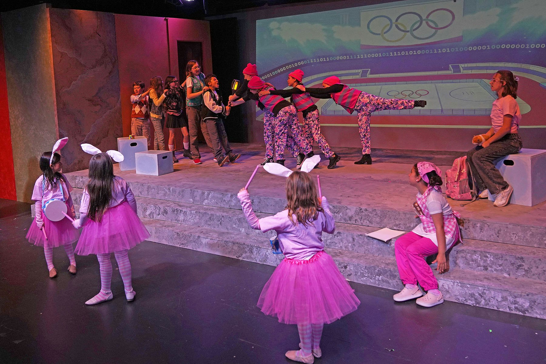 A lively stage scene with children performing in colorful costumes, some dressed as bunnies, set against an Olympic-themed backdrop.