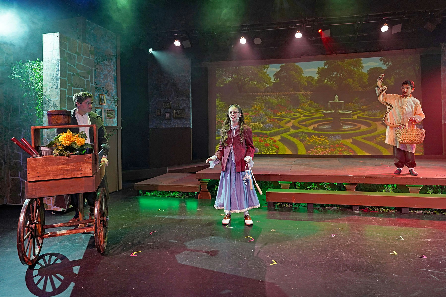 Three actors on stage in period costumes, with a floral cart and garden backdrop.