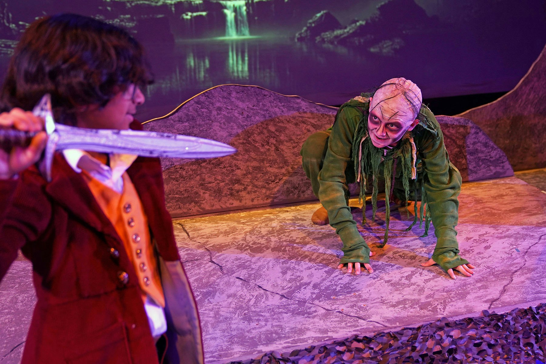 A person in a red coat holds a sword facing a costumed creature crouching onstage.